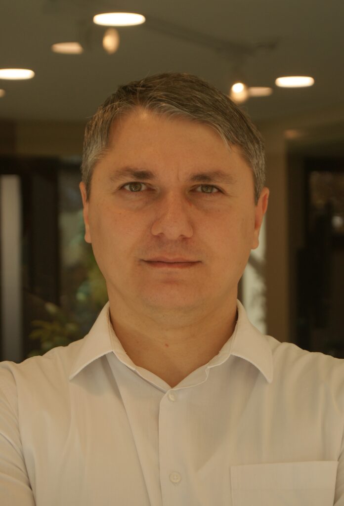 Portrait of Oleksandr Ganiev, Founder and CEO of ExImWheels, wearing a gray plaid suit and white shirt, smiling against a light gray background.